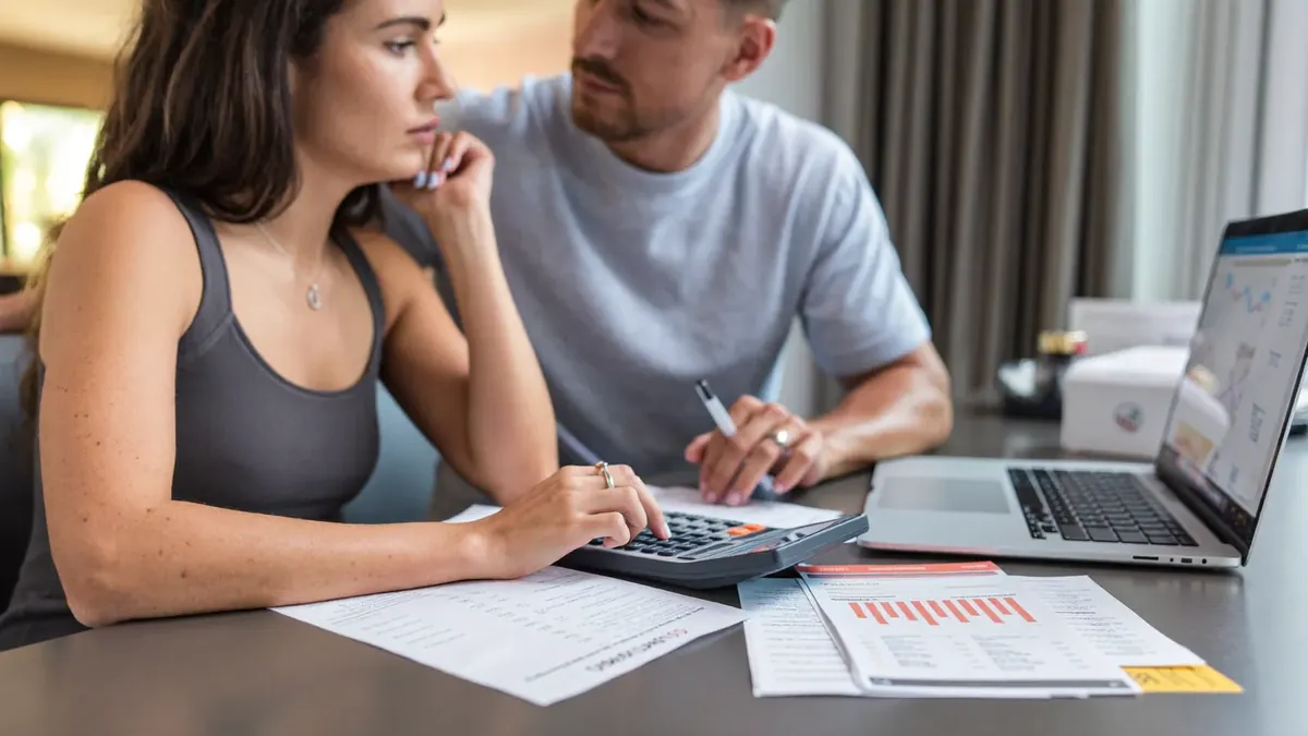 A couple in their thirties budgeting at a kitchen table with bills, a calculator, and a laptop showing health plan prices