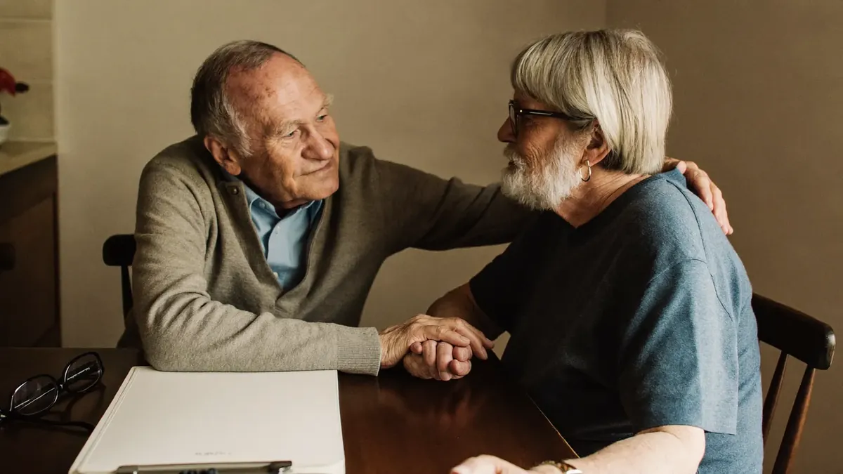 A retired couple holding hands reassuringly at their kitchen table