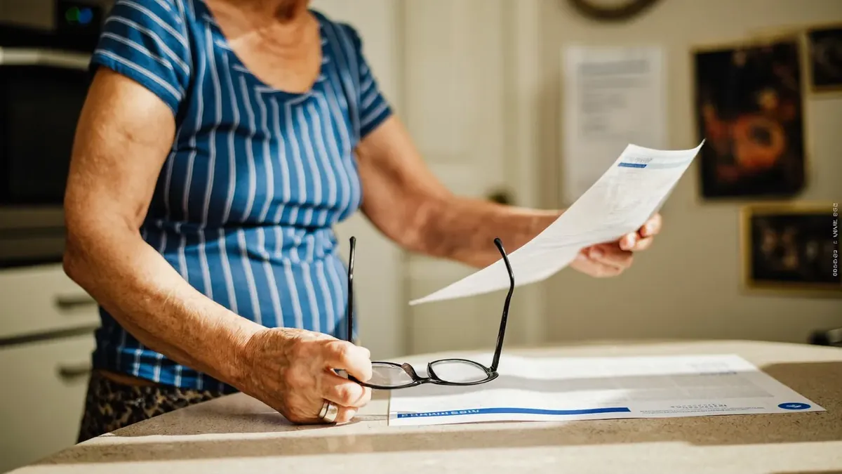 A 66 year old woman at her kitchen table opening a Medicare Part B premium letter, reading glasses on